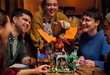 Promotional image of the Lego Ideas Dungeons & Dragons set Red Dragons Tale, showing a group of people at a table. Sitting on the table is the Lego set, showing a tavern, monsters, and a dragon.