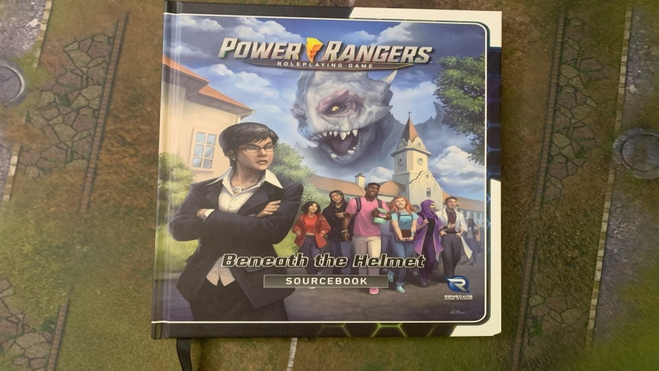 A copy of Power Rangers Beneath The Helmet sitting on a gaming mat.